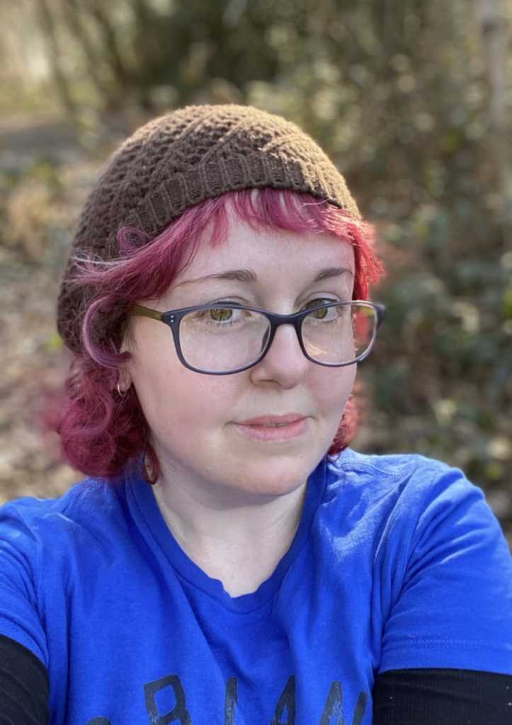 Photo of a white woman with curly hair dyed bright red, wearing glasses and a blue t-shirt. 